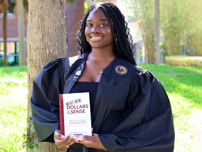 young woman in graduation attire holding Next Gen Dollars & Sense book