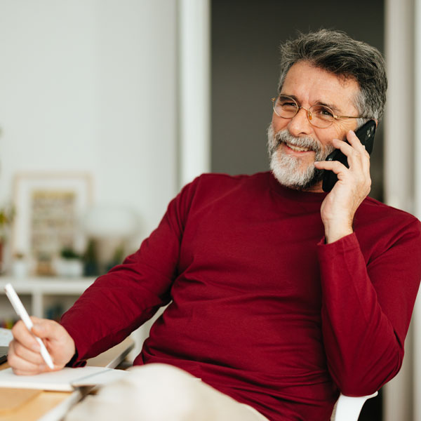 senior man speaking on the phone while sitting at home office desk writing notes in a notebook